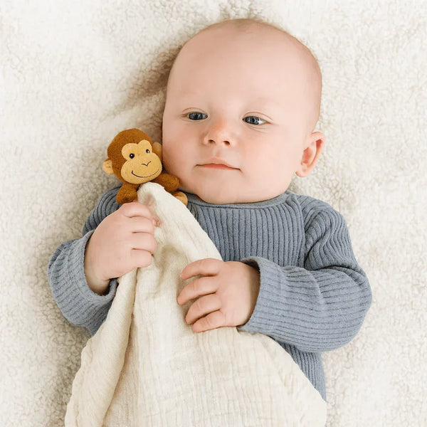 Baby holding cream cotton Me Lovey security blanket with brown monkey plush on soft white surface