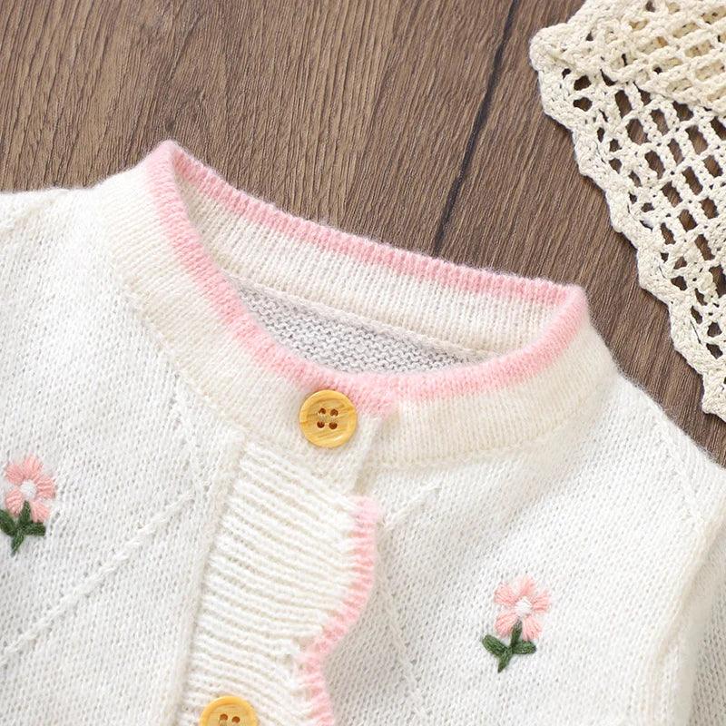 White baby knit romper with flower design in soft knit material featuring pink trim and wooden buttons