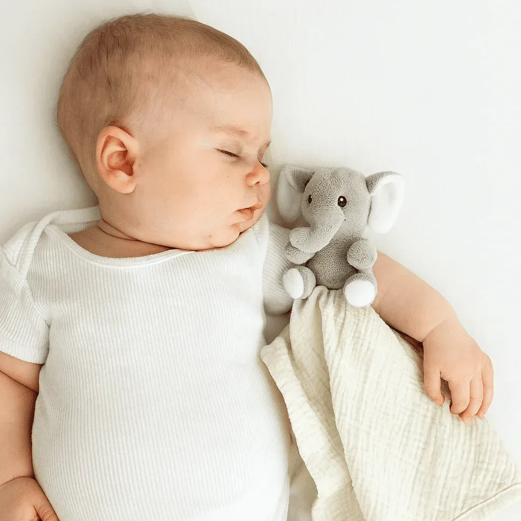 Gray plush elephant security blanket toy with cream muslin fabric held by sleeping baby in white onesie