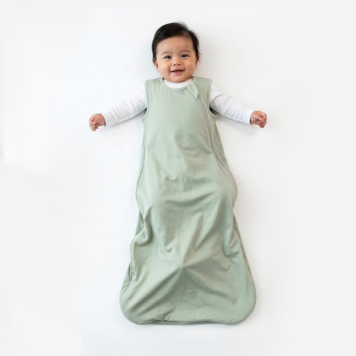 Infant wearing sage green sleep sack over white shirt lying on white surface
