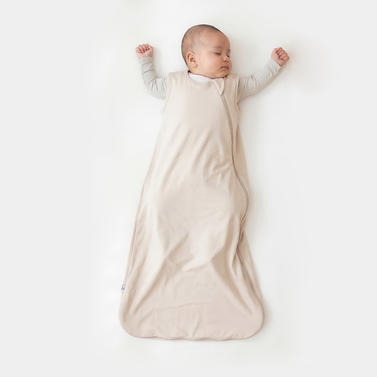 Baby sleeping in sand bamboo sleep sack over grey undershirt on white surface