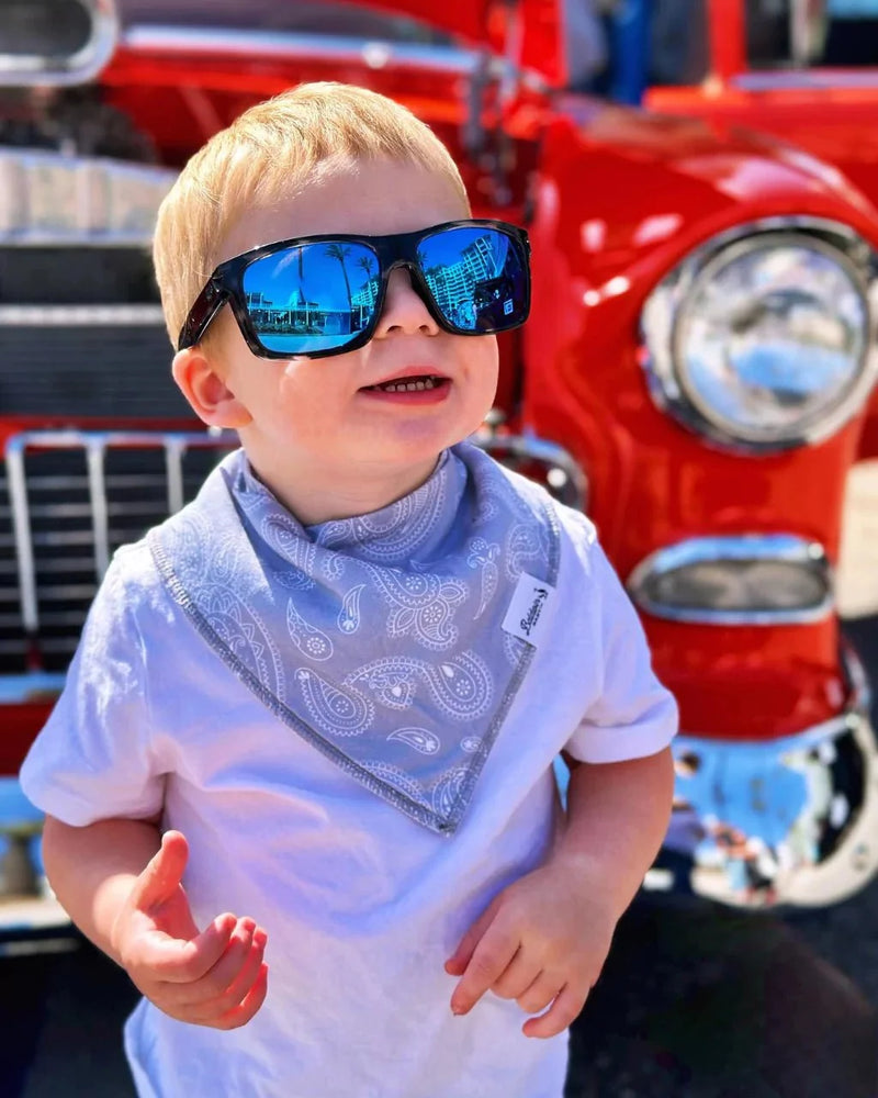 Baby wearing a blue paisley bandana bib and sunglasses in front of a red truck. Timeless Charm collection adds classic style and protection.