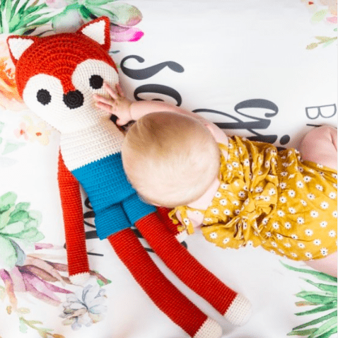 Baby in yellow dress hugging a large red, white, and blue crocheted fox toy with long limbs on floral bedding.