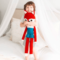Smiling child hugs a tall red and blue crocheted fox stuffed animal with long limbs and a pointed face in a cozy bed.