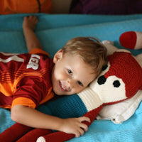 Smiling child hugging a large, long-limbed red and white knitted fox stuffed animal on a blue bed.