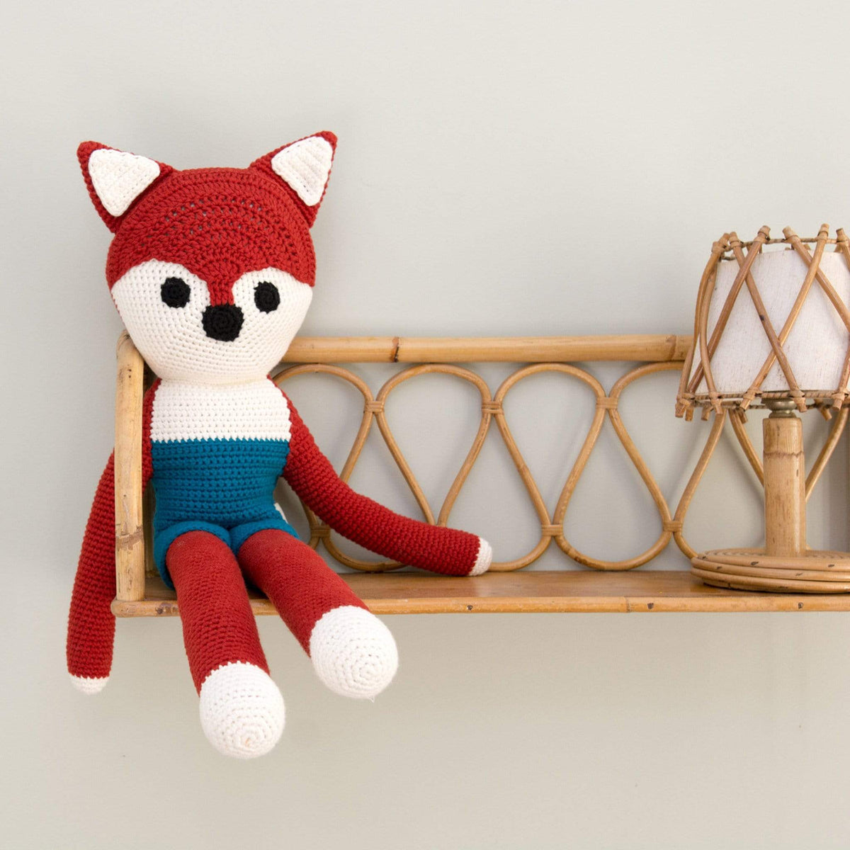 Hand-crocheted red fox stuffed animal with long limbs and a blue midsection sitting on a bamboo shelf.