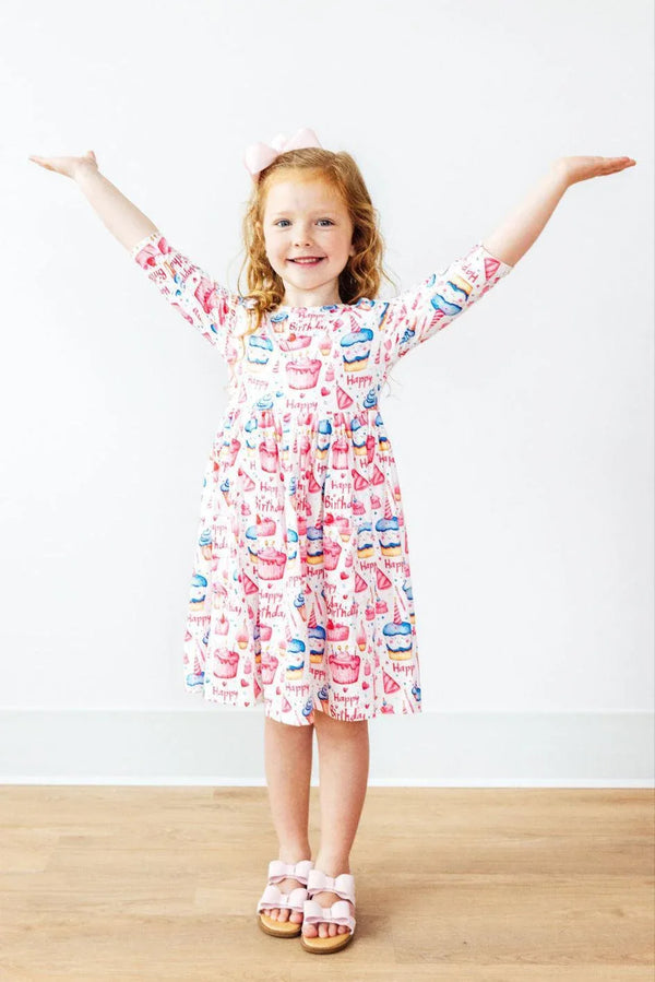 Young girl wearing white and pink birthday girl dress with cupcake print standing with arms raised