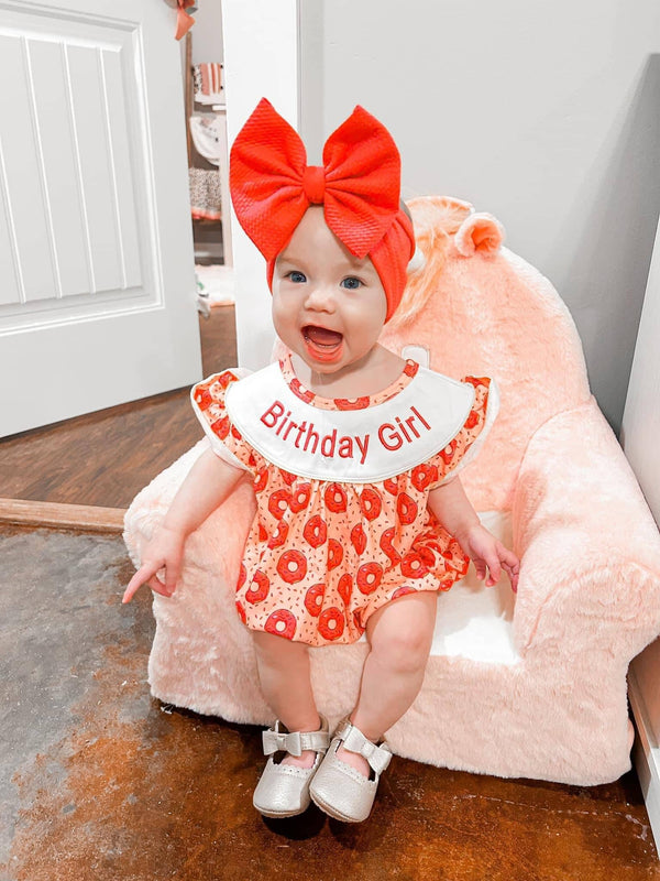 Red and white cotton birthday girl romper with donut print worn by baby girl sitting on pink plush chair