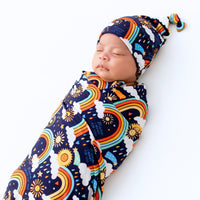 The SoftKnot Cap Here Comes the Sunshine (Blue) on a sleeping newborn, cozy bamboo hat with sun and rainbow print.