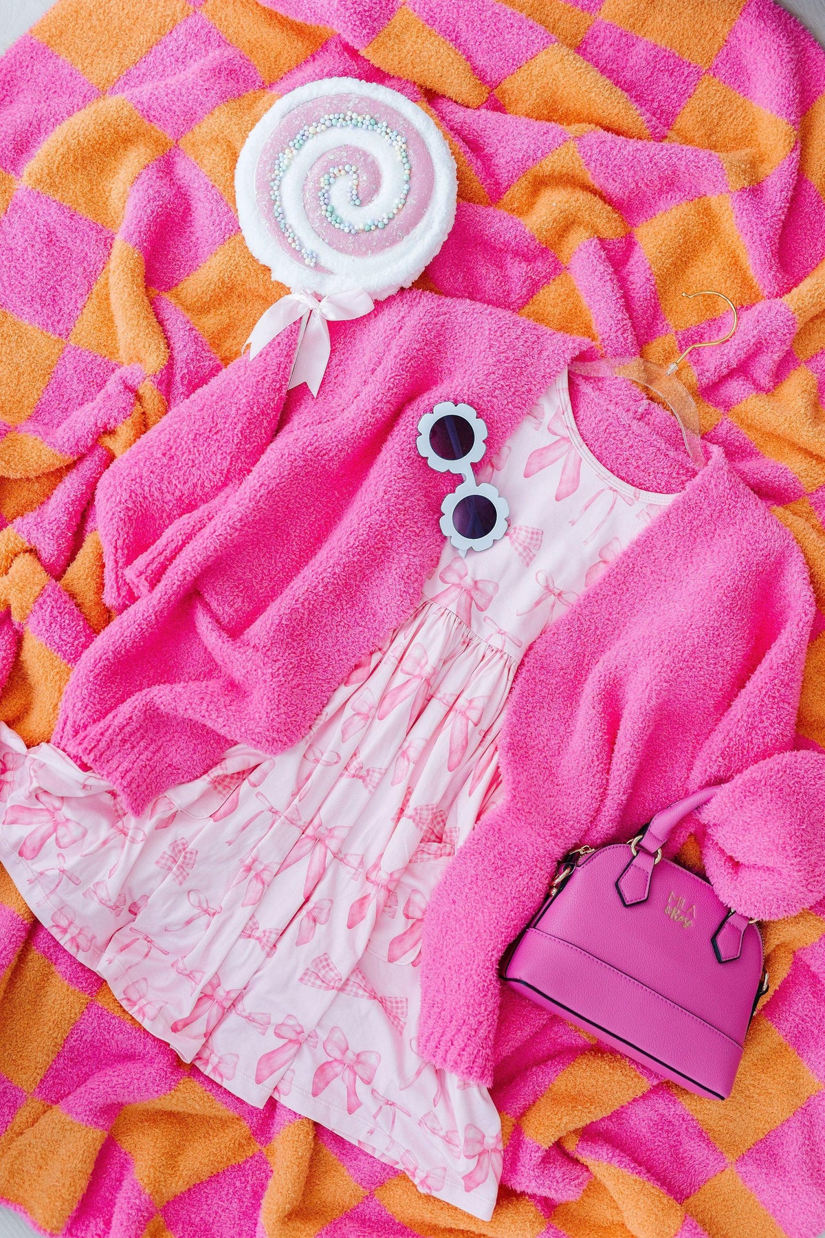 Pink blushing bows twirly dress for girls styled with a pink cardigan, sunglasses, and handbag on a colorful blanket