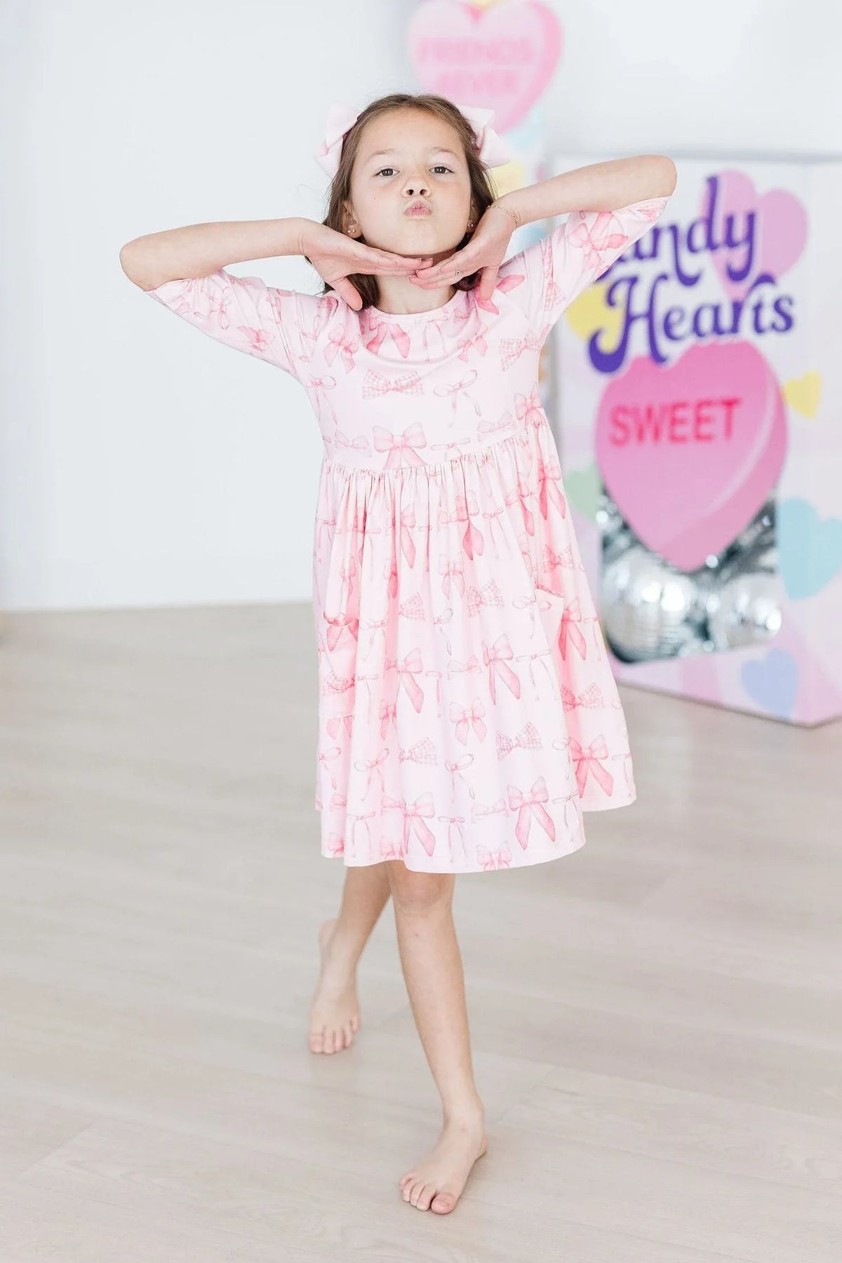 Young girl wearing a pink blushing bows twirly dress for girls posing with hands under chin barefoot