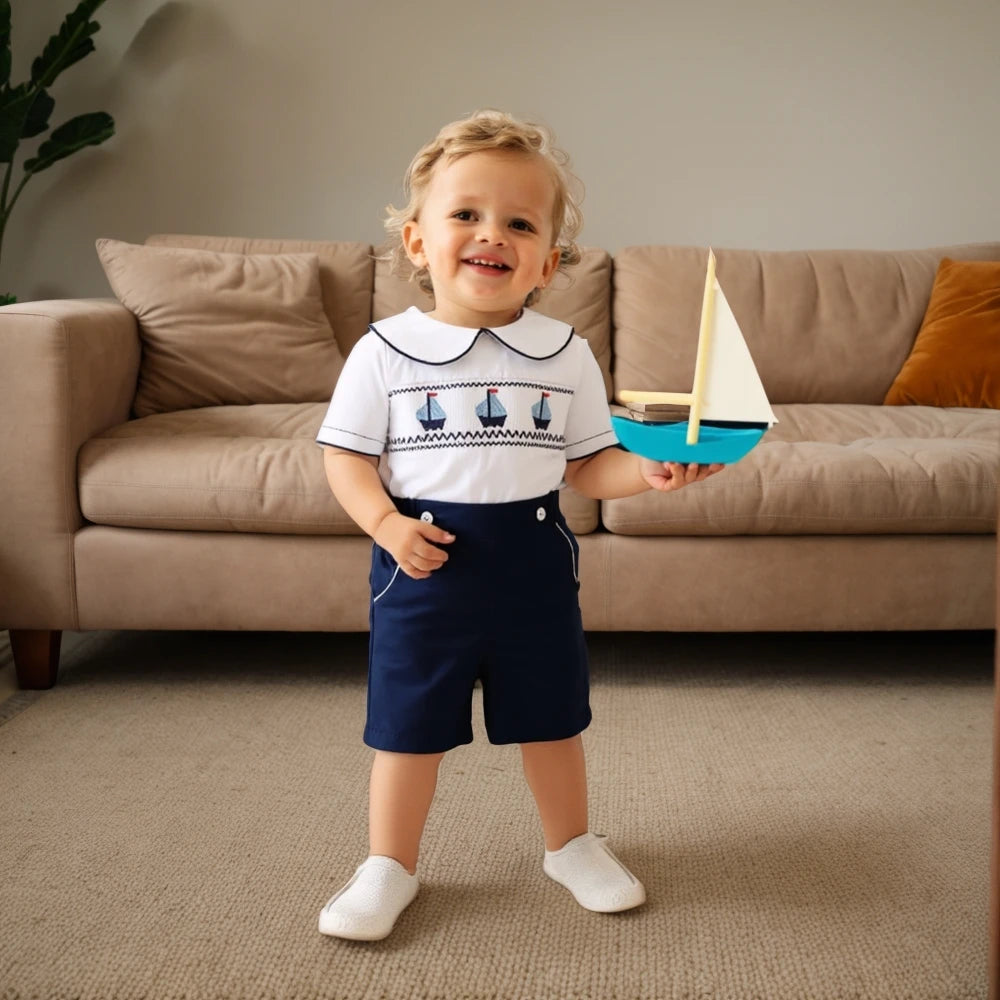 Toddler wearing a white cotton boys smocked sailboat outfit with navy shorts holding a toy sailboat