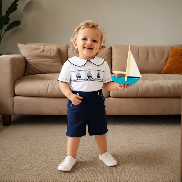 Toddler wearing a white cotton boys smocked sailboat outfit with navy shorts holding a toy sailboat