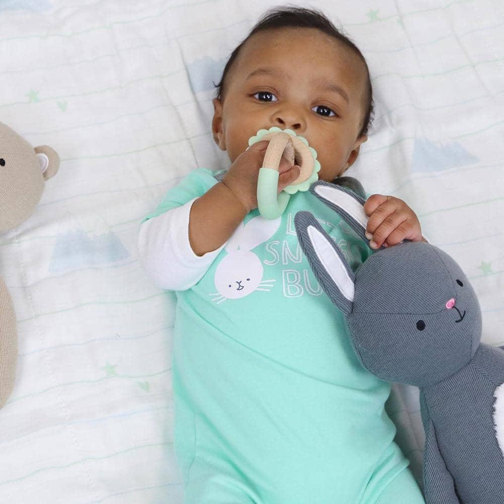 baby lying down holding gray bunny knit plush toy for baby while chewing a teether