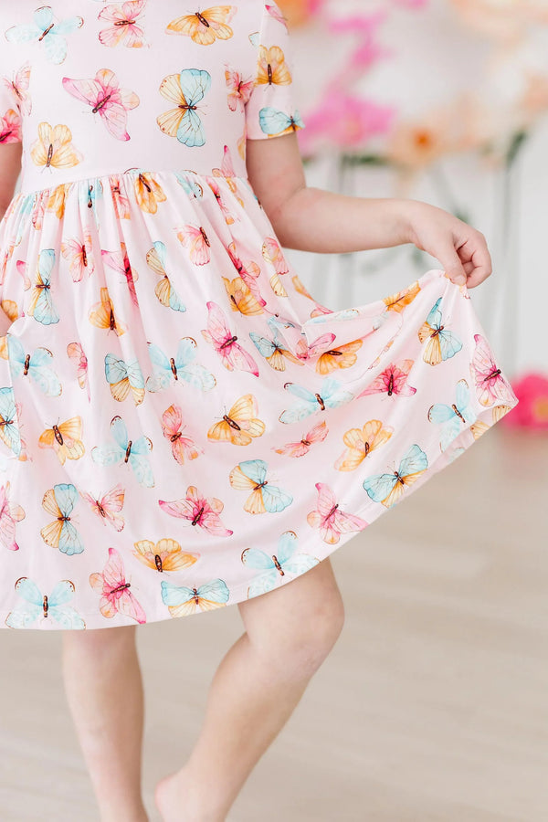 young girl wearing butterfly print twirl dress for girls in pastel pink holding skirt out