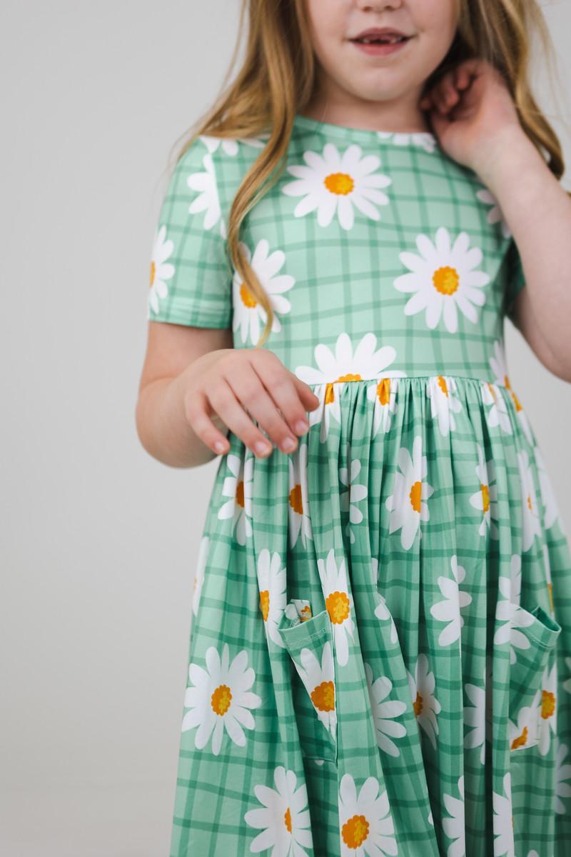 Close-up of a girl in the Off the Grid S/S Pocket Twirl Dress by Mila & Rose, highlighting the daisy pattern and handy pockets for all her treasures.