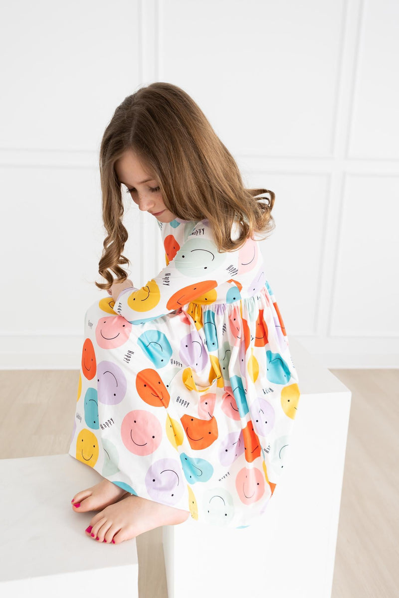 A little girl sitting gracefully in the 'Happy Days' 3/4 Pocket Fun Twirl Dress for Girls, highlighting its cheerful design and comfortable fit.