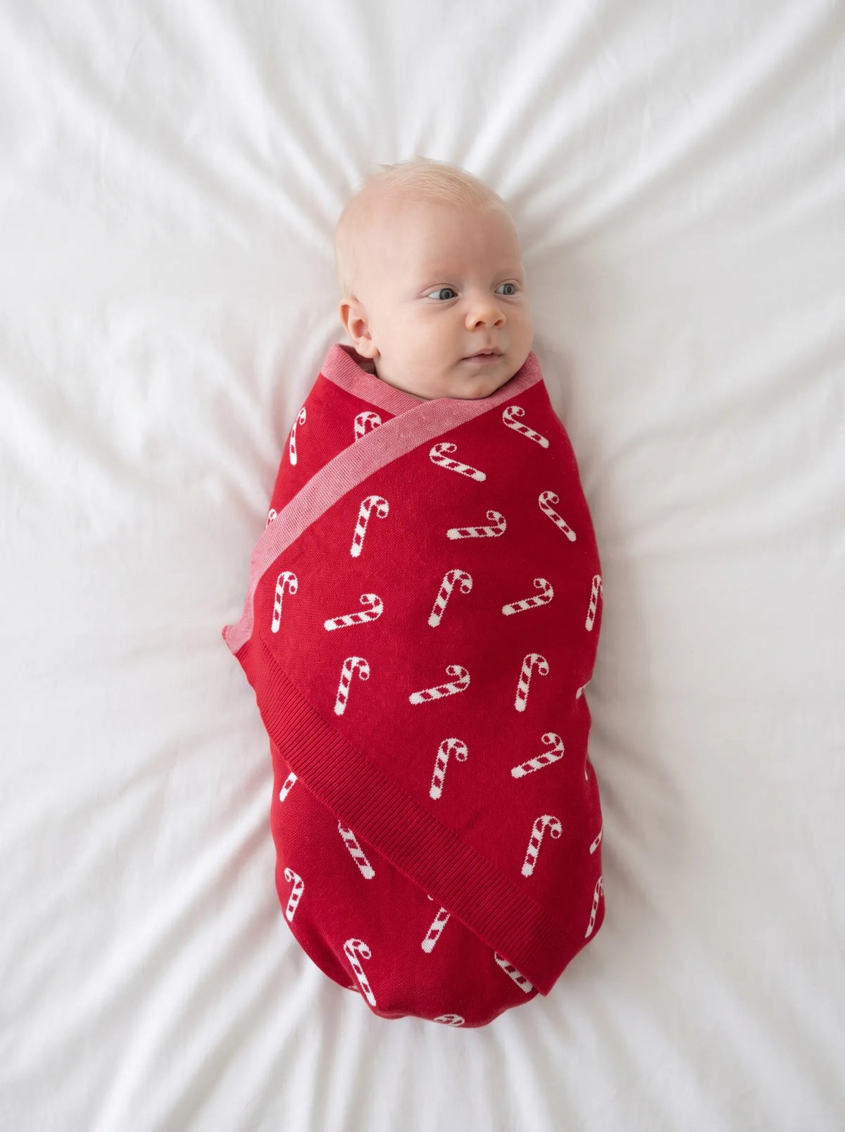 Newborn swaddled in red festive knit blanket on white bed sheet demonstrating size