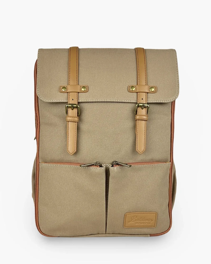 Beige rugged canvas diaper backpack with tan leather straps, brass buckles, and rust zipper trim on front pocket