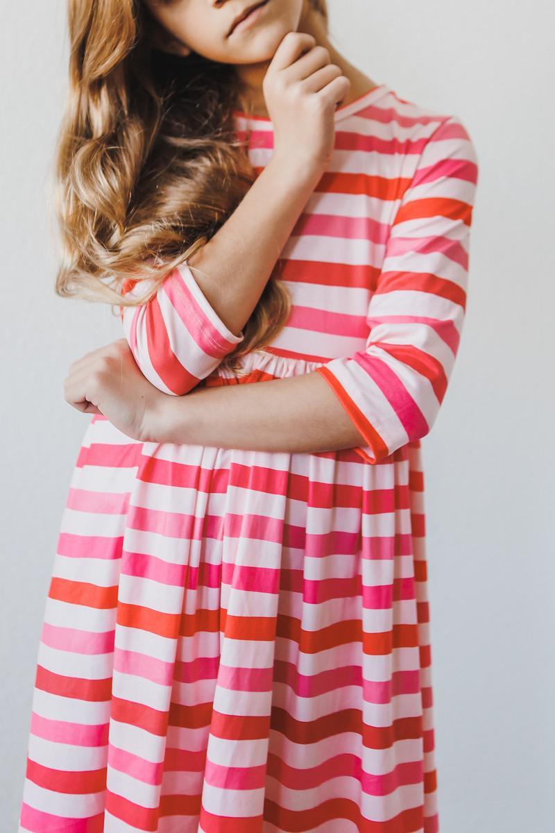 Close-up of a girl in the All You Need is Love 3/4 Sleeve Pocket Twirl Dress, showcasing the stylish pink and orange stripes, a great choice for a twirl dress for girls.