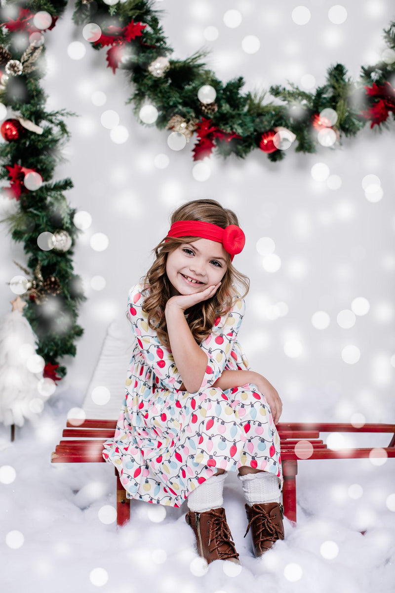 Smiling girl in the Merry & Bright 3/4 Sleeve Pocket Twirl Dress, sitting against a festive backdrop, embodying joy and holiday spirit.
