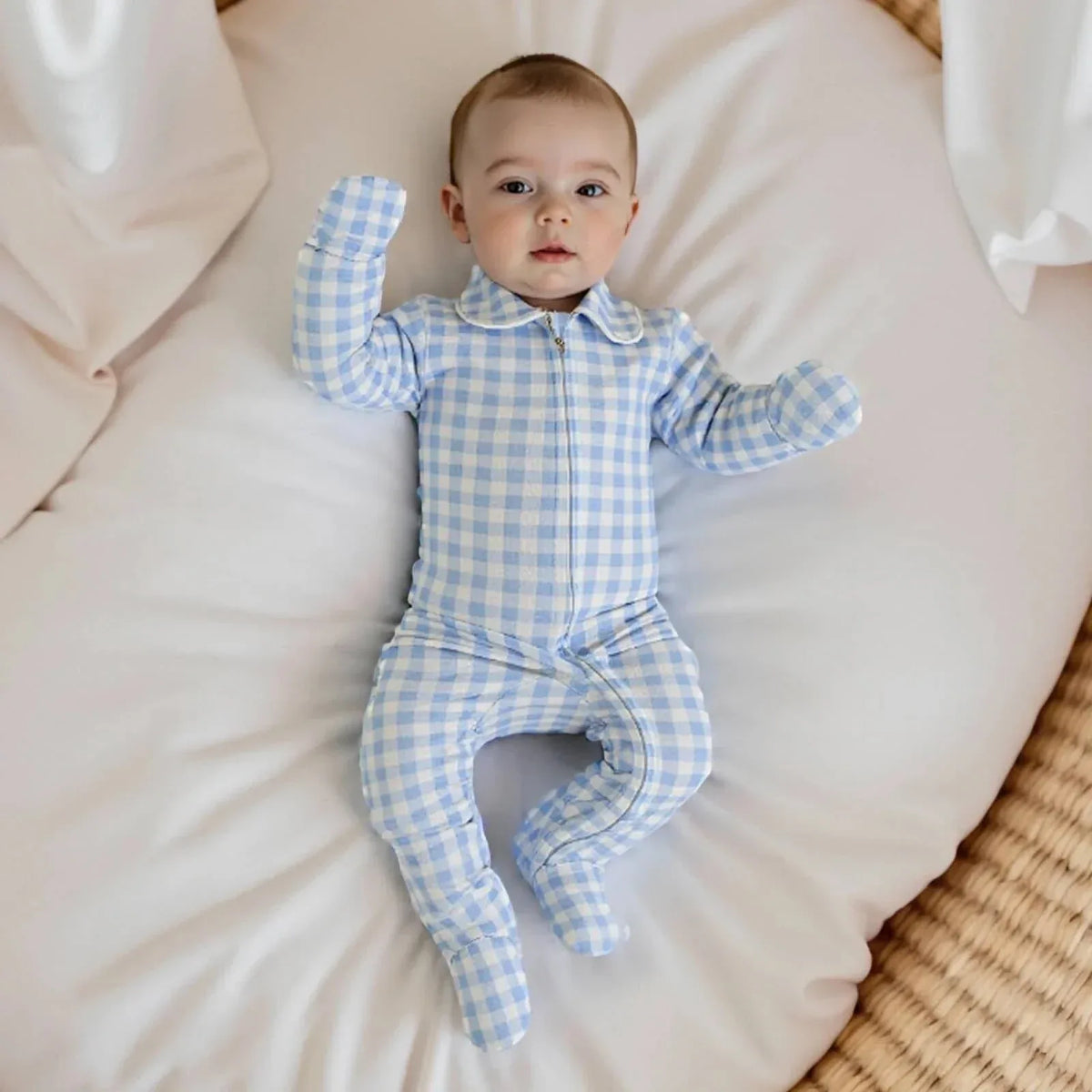 baby wearing blue and white checked baby zippered footie romper lying on a white cushion