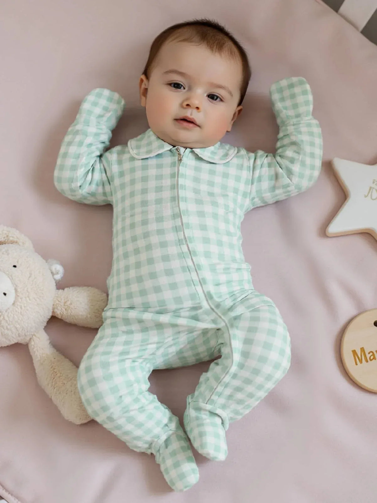 Baby wearing mint green checked baby zippered footie romper lying on a pink blanket with a stuffed bear