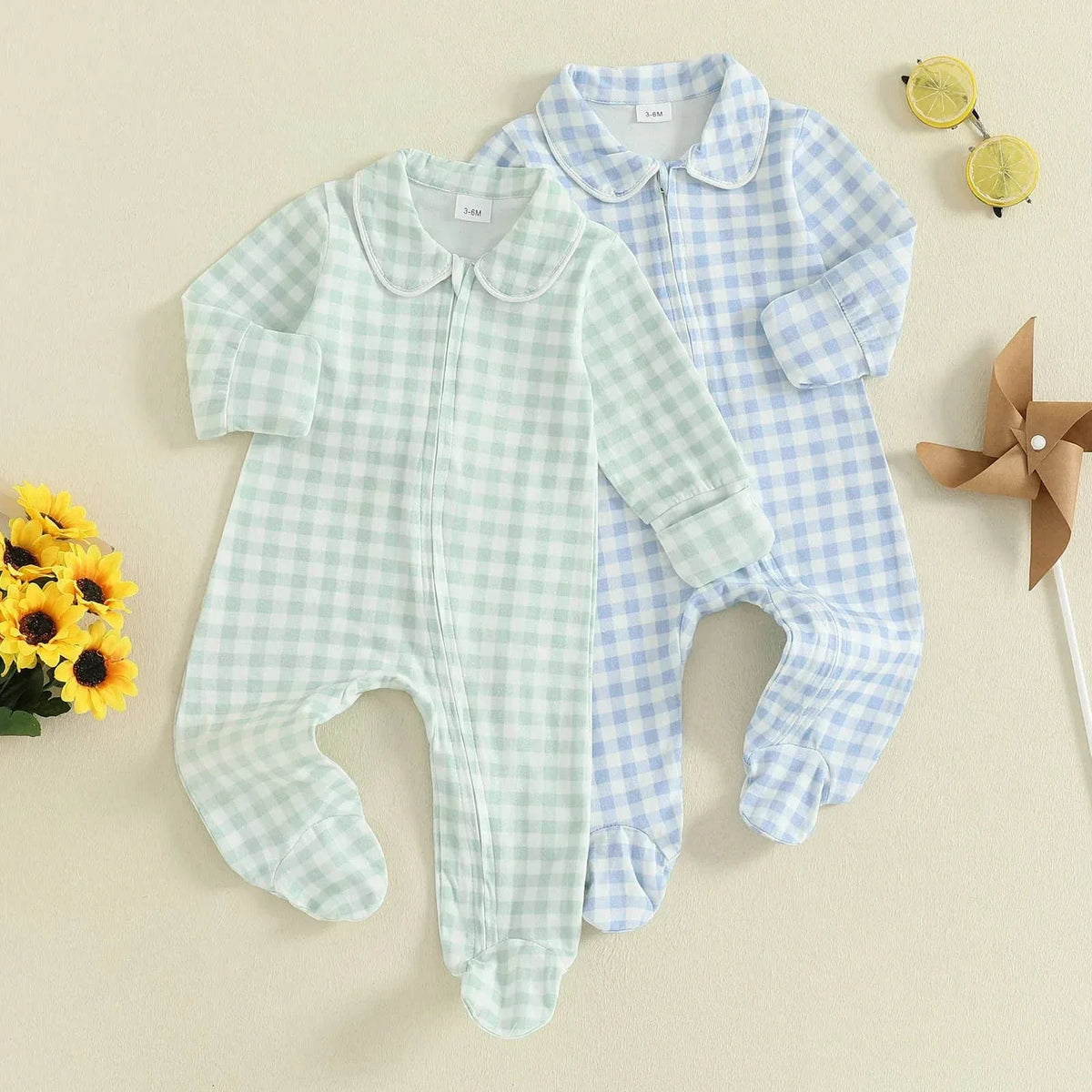Two checked baby zippered footie rompers in soft green and blue for infants laid flat with sunflowers and sunglasses nearby
