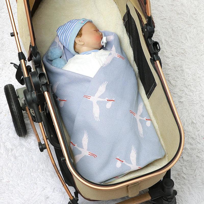 Baby boy wrapped in light blue children's knit blanket with white stork design in beige stroller