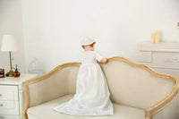White christening gown for boys toddler with cap, long flowing dress, standing on beige sofa