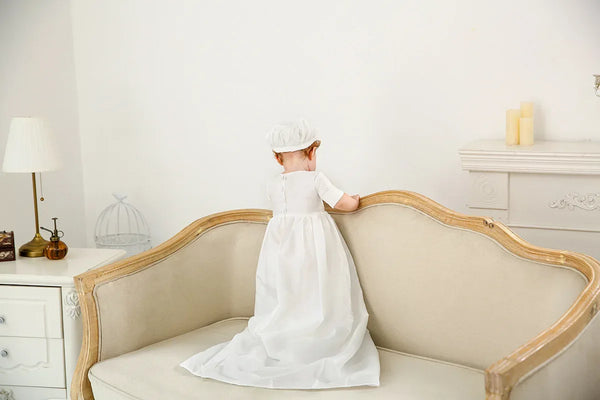 White christening gown for boys toddler with cap, long flowing dress, standing on beige sofa