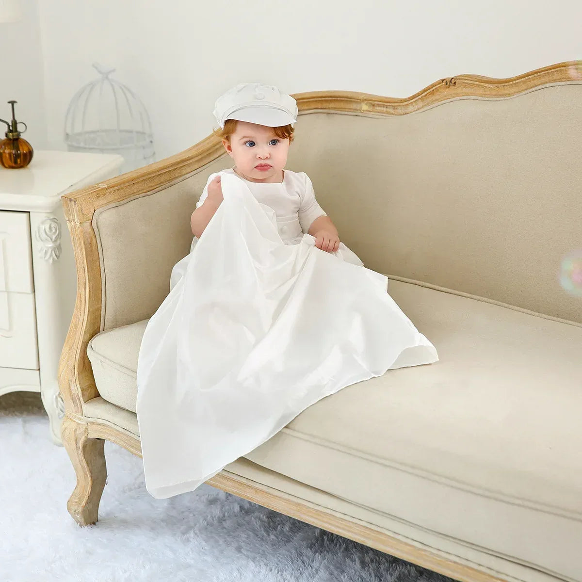 baby boy in white christening gown for boys with cap sitting on beige vintage sofa
