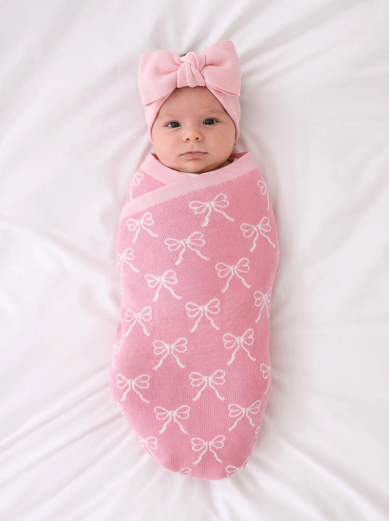 Newborn swaddled in pink bow knit blanket wearing matching headband on white textured sheet