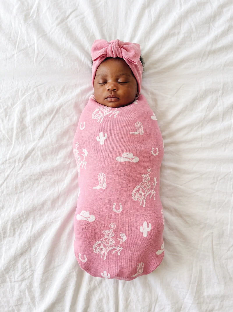 Sleeping newborn swaddled in pink cowgirl-themed knit blanket with matching bow headband on textured white sheet.