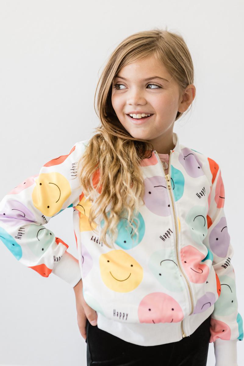 Girl smiling while wearing the 'Happy Days' Satin Jacket for Girls, adorned with colorful smiley faces, showcasing a stylish look with playful charm.