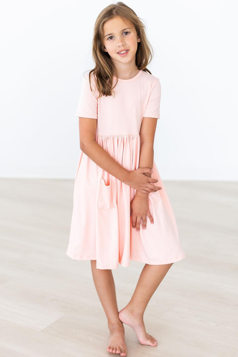 A confident girl posing in a Peach S/S Pocket Twirl Dress by Mila & Rose, demonstrating its comfortable fit and soft fabric.