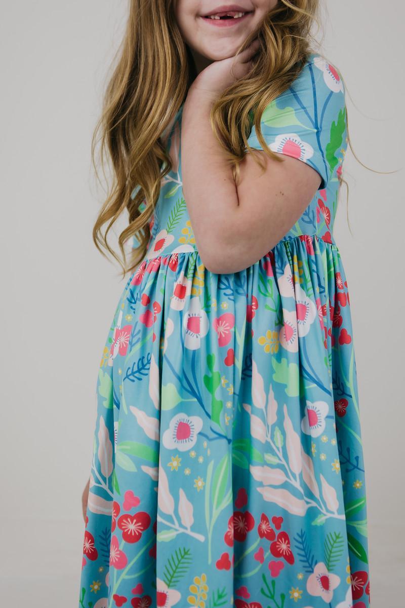 A happy child smiling while wearing the Spring Breeze S/S Pocket Twirl Dress, highlighting the playful full skirt and bright floral print, ideal for twirling and fun activities.