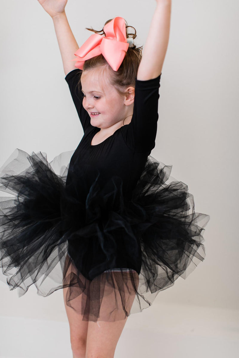A happy child wearing a Black 3/4 Tutu Leotard by Mila & Rose, celebrating with arms raised, accentuated by a pink bow and fluffy tutu.