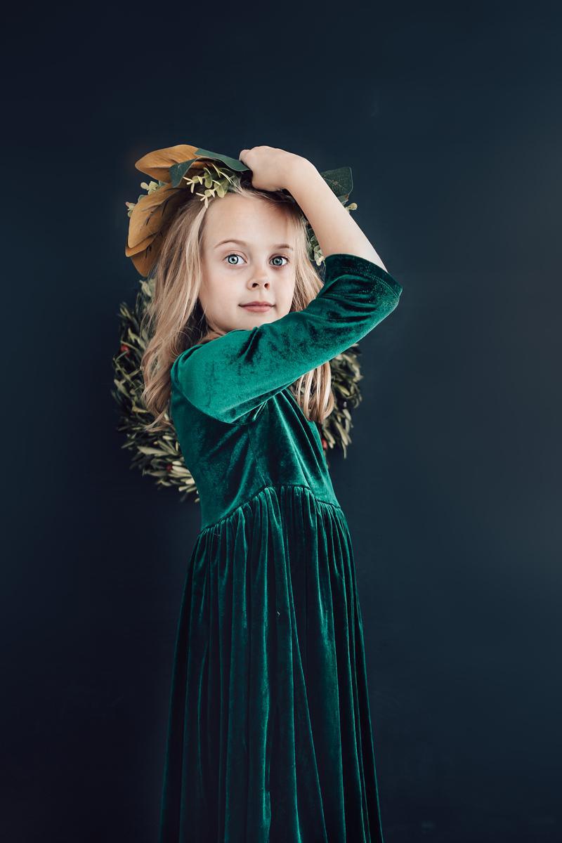 Girl with flowing blonde hair wearing a Hunter Green Velvet Twirl Dress by Mila & Rose, holding a floral crown against a dark background.