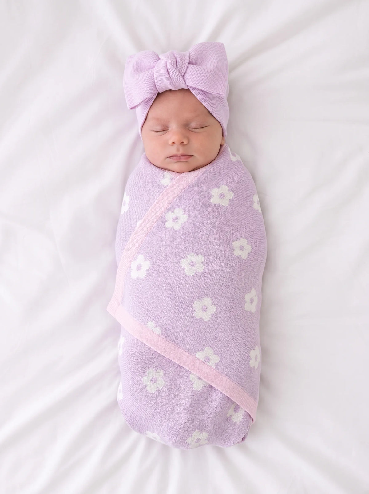 Newborn swaddled in lavender-pink daisy jacquard knit baby blanket with white flowers and pink trim, wearing a matching bow