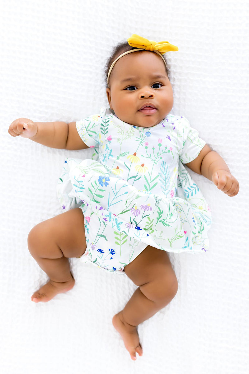 Baby in a Spring Dandelion Floral S/S Twirl Bodysuit by Mila & Rose, showcasing comfort and style with a cheerful floral design and a cute yellow bow headband.