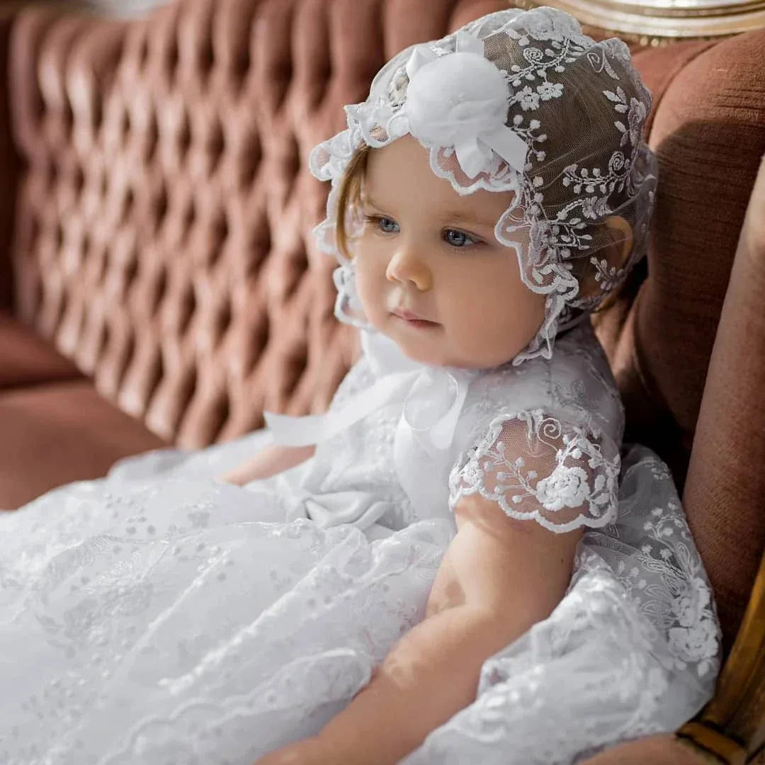 baby girl wearing delicate lace baptism gown for baby girls in white sitting on brown sofa