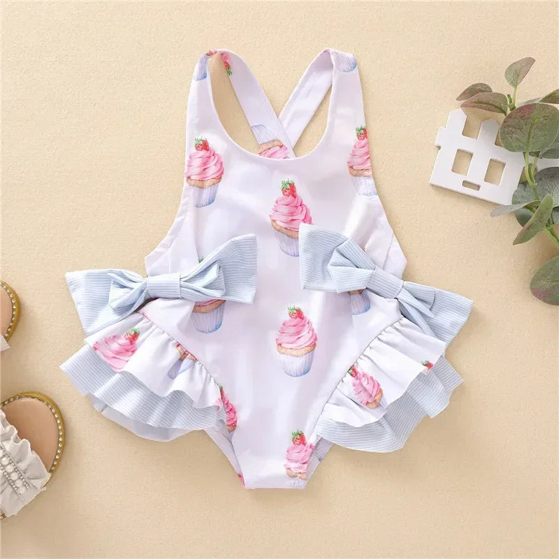White dessert themed baby swimsuit with pink cupcake print and blue bow ruffle details for infants