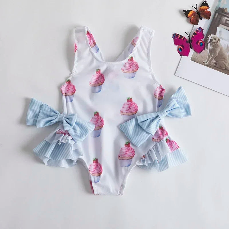 White dessert themed baby swimsuit with pink cupcake print and blue bow details for infants