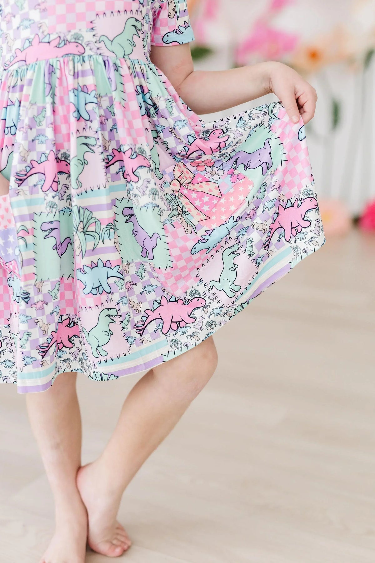 young girl wearing pastel pink dinosaur twirl dress for girls holding skirt while standing barefoot indoors