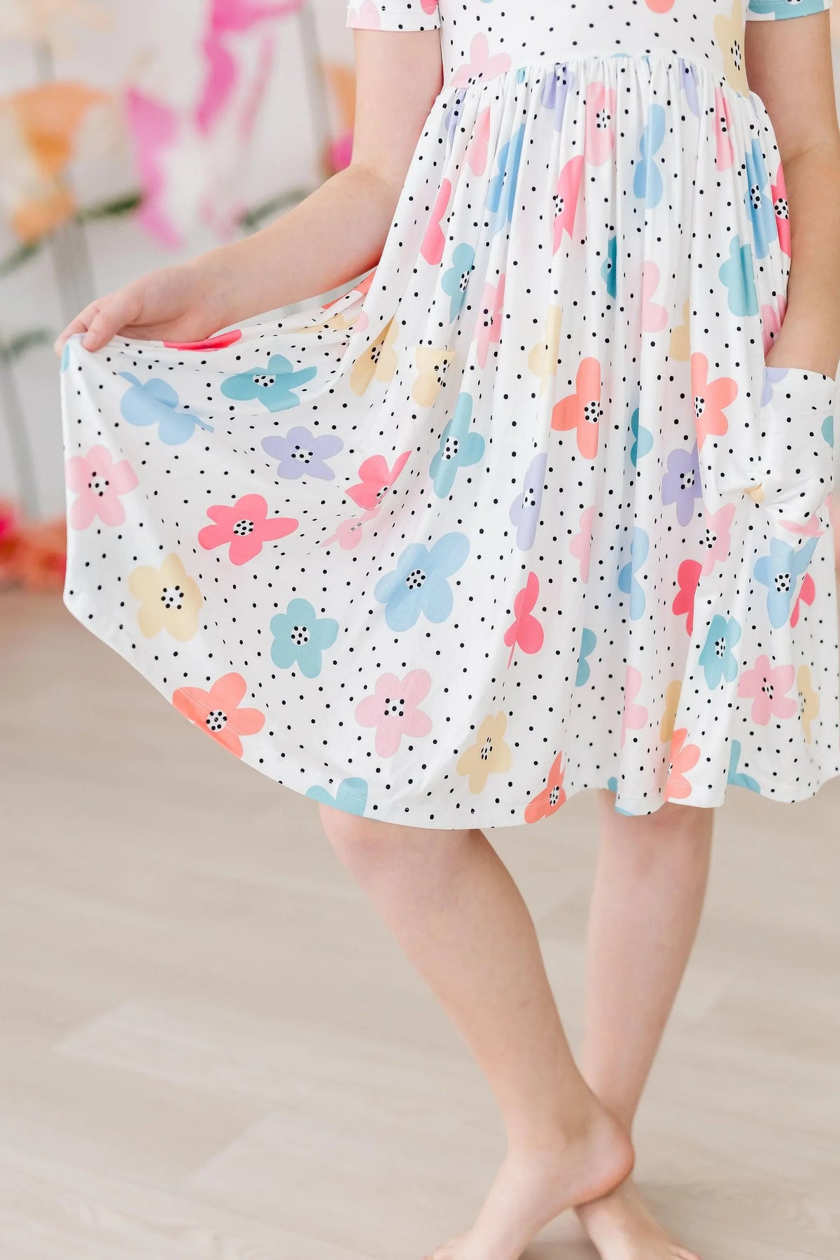 Young girl wearing white dots and daisies pocket dress for girls holding skirt while standing barefoot