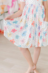 Young girl wearing white dots and daisies pocket dress for girls holding skirt while standing barefoot