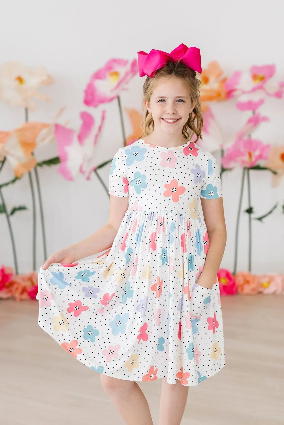 young girl wearing white dots and daisies pocket dress for girls with pink bow smiling and holding dress
