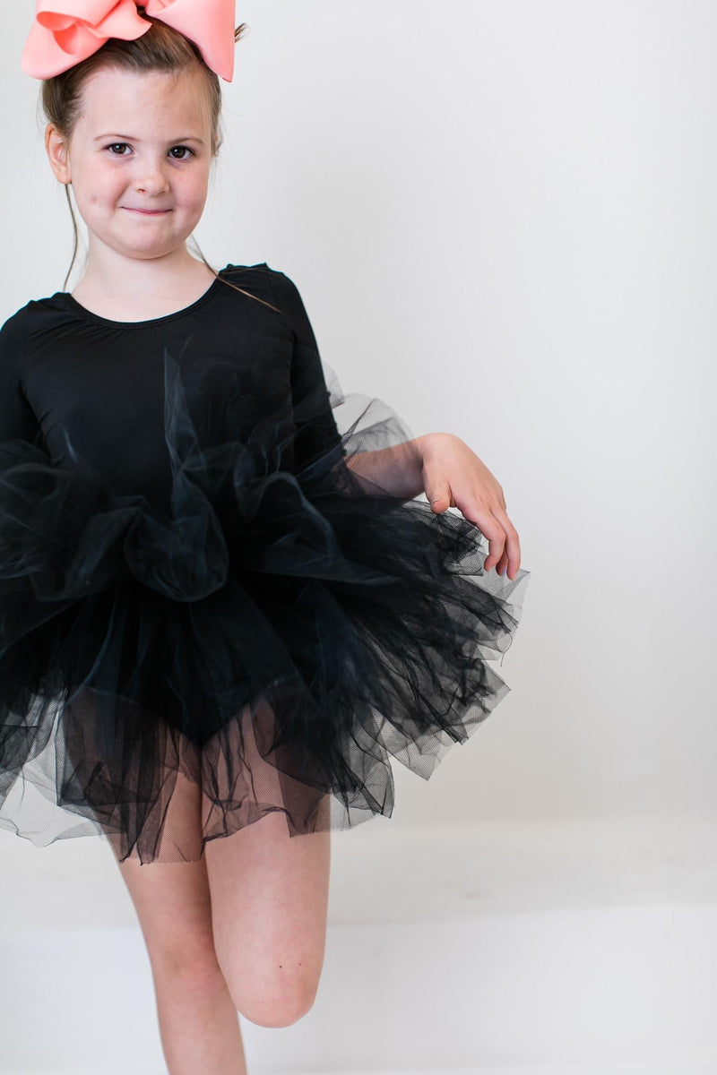 Young girl wearing a Black 3/4 Tutu Leotard with fluffy layers, showcasing a playful pose and a big pink bow in her hair.
