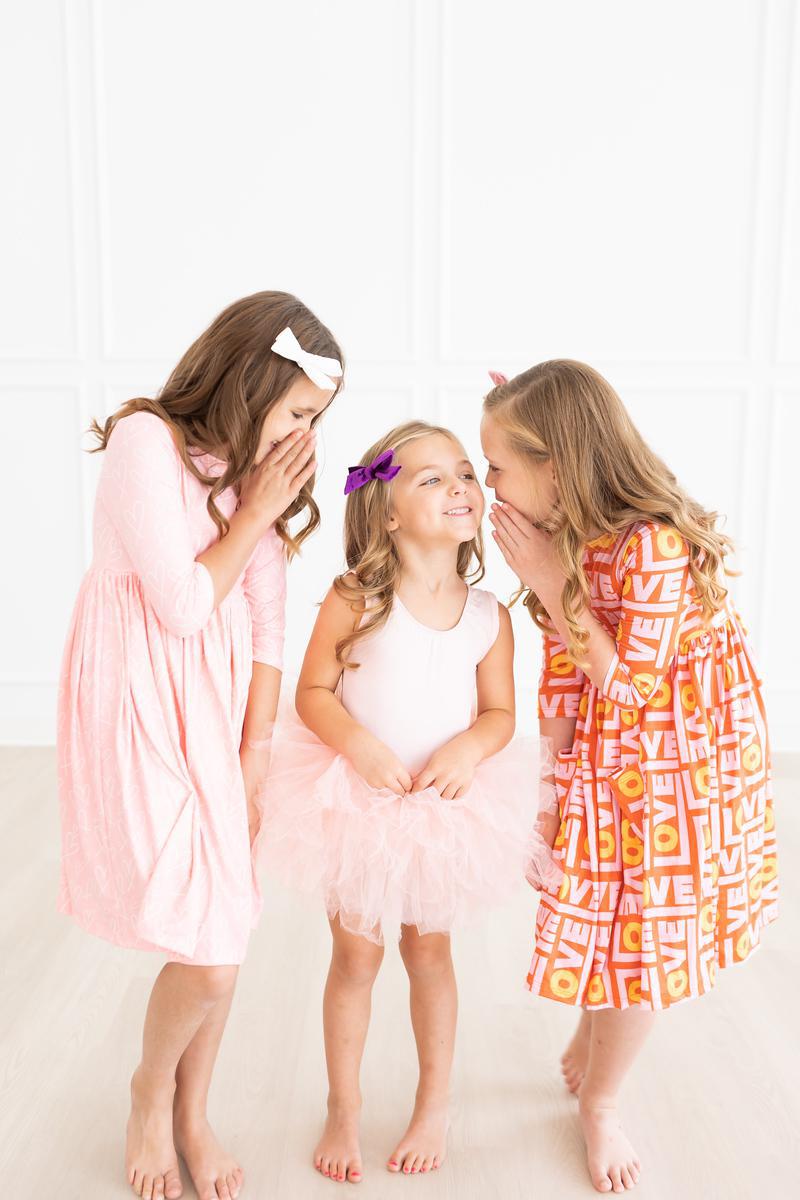 Three girls sharing giggles, one dressed in the 'Elana' Ballet Pink Tank Tutu Leotard, radiating joy at a fun gathering.